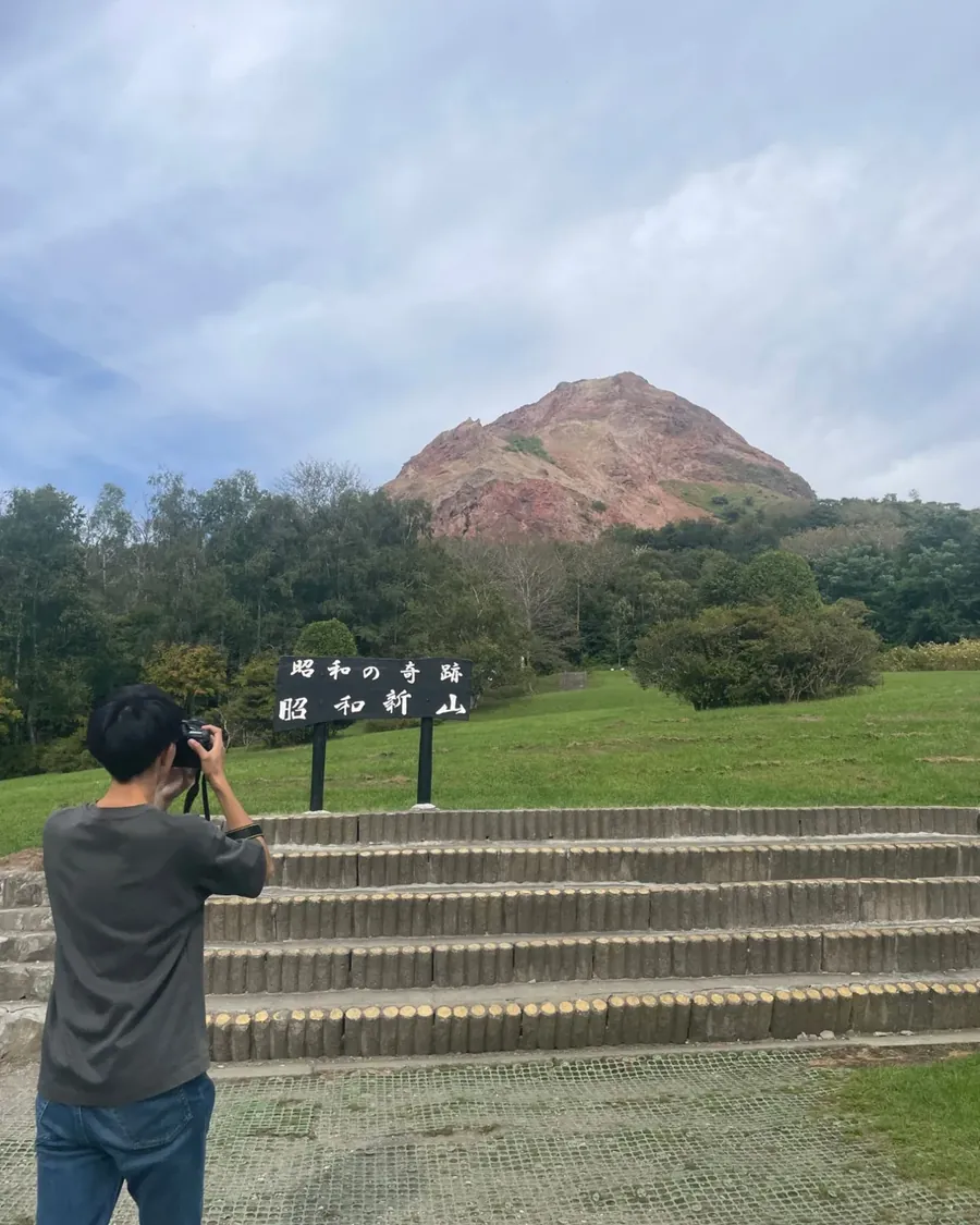 昭和新山の赤い溶岩ドームと「昭和の奇跡 昭和新山」の札。手前でカメラを構える人の後ろ姿。